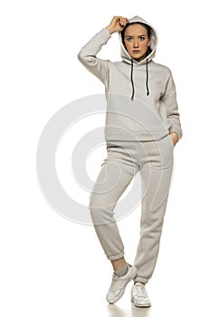 Front view of a young woman in a gray tracksuit and hood posing to a white background in the studio