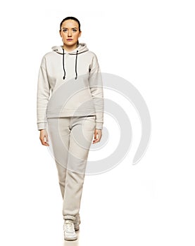 Front view of a young woman in a gray tracksuit and hood walking to a white background in the studio