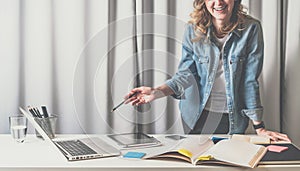 Front view of young woman standing in designer office near table on which there are catalogs,laptop,tablet computer.