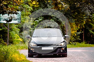 Frontal photo of a black automobile rides on the road in the forest with headlights on and no car number at the front of