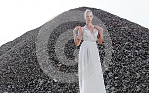 Frontal portrait of blonde woman with hair tight up in white bride dress, in day time, over piles of stones. Copy space