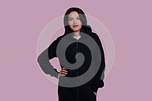 Frontal portrait of a cheerful brunette female model dressed in a black swater with hood, isolated on pink background.