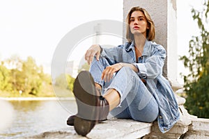 Frontal portrait of a single pensive teen girl, dressed in denim and boots, seated outside near river, looking away.