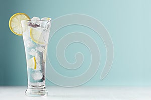 Frosty transparent lemonade with lemon slices, ice cubes and mineral water in elegant glass on white wood table, mint color wall.