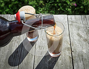 Frothy dark beer pouring into tall glasses from a brown glass bottle in summer garden on rustic wooden table