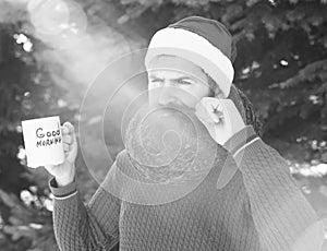 Frown handsome man in santa claus hat, bearded hipster with beard twirls moustache and drinks from cup with good morning