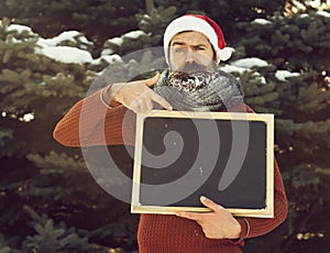 Frown man in santa claus hat, hipster with beard and moustache covered with white frost, points at blank black board or