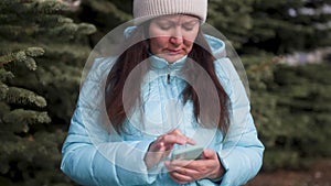 Frown woman typing message chatting in smartphone outdoors in fir tree park.