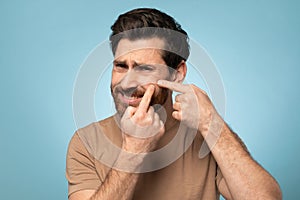 Frowning middle aged man popping inflamed pimple on face, looking at camera while standing on blue background