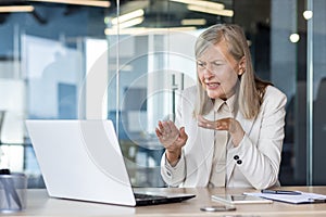 Frowning senior woman hosting online video call by laptop and gesturing actively with hands in glassy office. Stressed