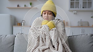 Frozen Asian chinese woman in warm hat and mittens rubbing freezing hands sit in cold kitchen frosty temperature heating