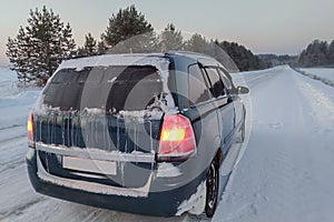 A frozen car stands on the side of a slippery winter road at dusk. Orange warning lights are on. The rear window was covered with