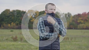 Frozen caucasian man standing on the autumn meadow and zipping his vest. Bearded guy with grey hair waiting for someone