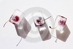 Frozen cherries in ice cubes on white textured background. Hard sunlight, dark shadow, flat lay. Preservation of summer vitamins