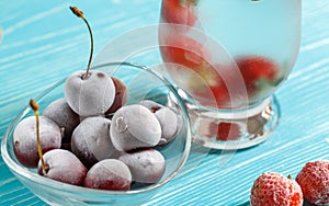 Frozen cherries and strawberries in a glass bowl and a glass of cold water. Close-up on a blue wooden background