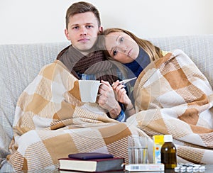 Frozen couple sitting on couch under blanket with thermometer