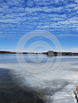 Frozen lake or sea melting in the spring. Blue sky with unusually shaped soft clouds and forested islands in the distance. Finnish