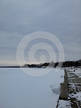 Frozen lake with a thick layer of snow. Small urban forest with bare trees.