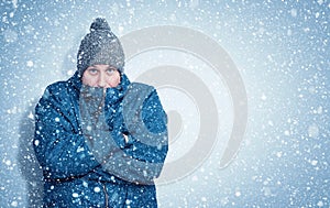 Frozen man in a blue jacket and hat stands against the wall, snow is falling around