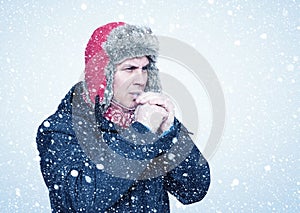 Frozen man in a jacket and a hat warms his hands, it is snowing around