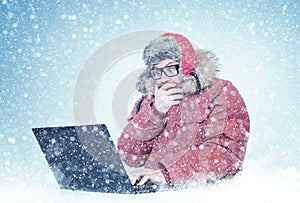 Frozen man in red winter clothes and glasses working on a laptop in the snow. Cold, frost, blizzard, computer
