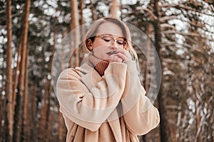 Frozen mid age woman in winter forest warming up hands outdoors looking down. Blonde girl in coat sweater and eyeglasses