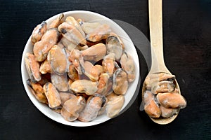 Frozen, peeled mussels, without shells, are in a round salad bowl, next to which there is a wooden spoon filled with mussels. dark