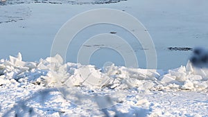 A frozen river bank with fragments of ice floes with blurred branches in the foreground