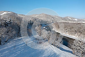 A frozen river flows between trees covered with white snow. On a frosty sunny December day in a forest covered with