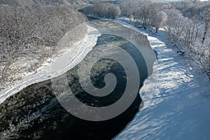 A frozen river flows between trees covered with white snow. On a frosty sunny December day in a forest covered with