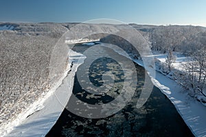 A frozen river flows between trees covered with white snow. On a frosty sunny December day in a forest covered with