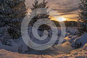 Frozen river at sunset surrounded by snow and evergreen trees.