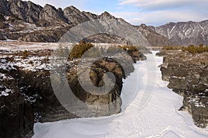 Frozen riverbed squeezed into rocky high banks flowing through a snow-covered valley surrounded by mountain ranges in winter