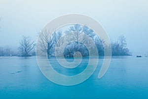 Frozen turquoise lake in misty winter morning, with trees without leaves