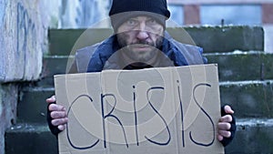 Frozen untidy young man sits on the steps and holds a handwritten CRISIS poster.