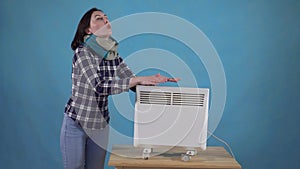 Frozen woman in scarf next heated with an electric heater on a blue background