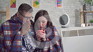 Frozen young woman and man wrapped in a blanket in the living room are heated next to electric heaters close up