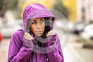 A frozen young woman in a rain jacket has a hood on and protects her head from the rain and wind during late autumn
