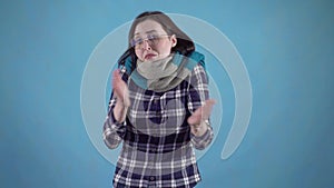 Frozen young woman in scarf and glasses can`t get warm standing on blue background