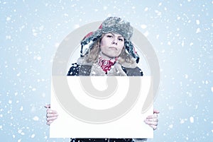 Frozen young woman in winter clothes holds an empty white poster in her hands, it is snowing on a blue background.