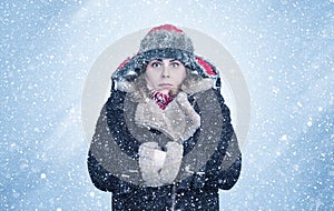 Frozen young woman in winter clothes warms her hands in the sleeves of a sheepskin coat, it`s snowing around on a blue background
