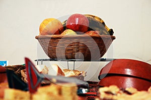A fruit casket with apple, banana and orange standing on a metal table with loads of other kitchen gear in front of it