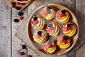 Fruit dessert tartlets with vanilla custard and fresh raspberries, blackberry, peach. Top view. Dark rustic style.