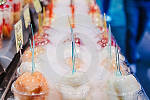 Fruit juice mix with ice arranged in plastic cups on a market stall with small fork inside, takeaway snack