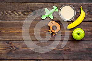 Fruit puree for baby. Jar with food, apple, banana, toys on dark wooden background top view copy space