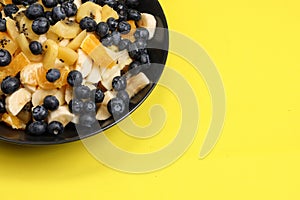 Fruit salad with blueberries, banana, orange, apple and kiwi on a black plate isolated on a yellow background. Vitamin cocktail.