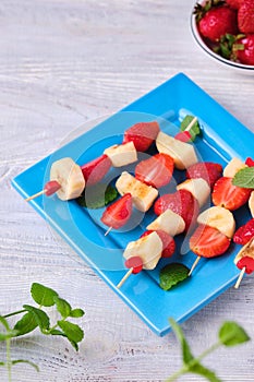 Fruit skewers with strawberry and banana on a blue square plate on a light wooden background. Dishes party, children, holiday