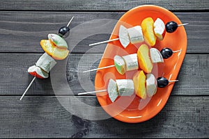 Fruit slices strung on a wooden stick in the form of a kebab in an orange saucer, on a wooden background.