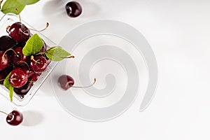 Fruit summer snack - fresh wet ripe red cherries with green leaves in transparent box package in soft light white wood board.