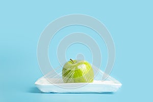 Fruits in plastic packaging from the supermarket are minimal. Apple in cellophane and non-degradable plastic on a blue